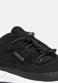 Zapatilla negra con parte superior de ante texturizado, cordones elásticos y un cierre de plástico. Suela de goma con un patrón de banda de rodadura distintivo. Logotipo de Timberland visible.