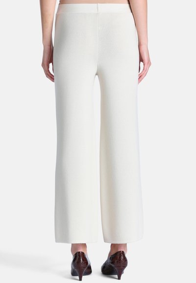 Personne portant un pantalon blanc taille haute à jambes larges et des chaussures marron foncé à talons bas, debout devant un fond uni.