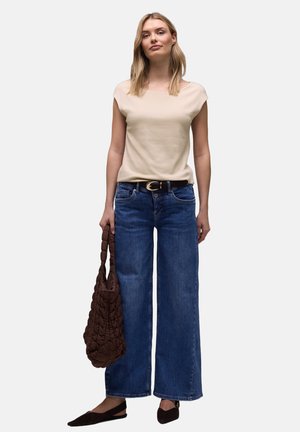 Joven mujer con camiseta beige de manga corta, pantalones vaqueros azules de pierna ancha, cinturón negro, zapatos planos negros con punta y un gran bolso marrón tejido.