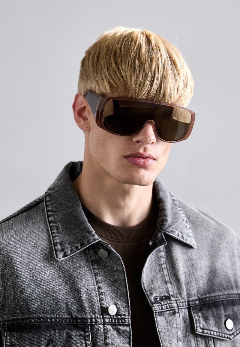 Übergroße braune Sonnenbrille mit dunklen Gläsern, rechteckiger Form. Getragen mit einer grauen Jeansjacke und einem braunen strukturierten Pullover darunter.