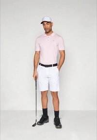 Polo rosa claro de manga curta, calções brancos, cinto preto e sapatos de golfe pretos. Segurando um taco de golfe, usando um boné branco.