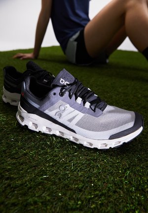 Chaussures de sport grises et noires sur gazon artificiel, avec une personne assise en arrière-plan, portant un short et un t-shirt bleu marine.
