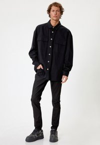 Musta nappipaita, jossa on kaksi rintataskua, käytetty slim-fit mustien housujen ja mustien teksturoidun tennarien kanssa. Malli seisoo käsi taskussa.