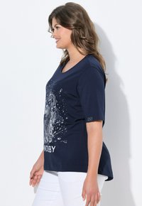 Hymyilevä nainen, jolla on laineikkaat hiukset, pukeutuneena tummansiniharmaaseen T-paitaan, jossa on koirakuva ja valkoista tekstiä, yhdistettynä valkoisiin housuihin, tasaisen taustan edessä.