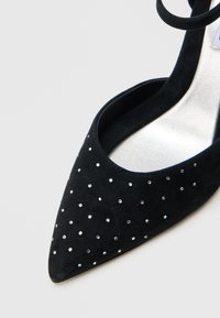 Μαύρο σουέντ παπούτσι με μυτερή μύτη, εσωτερικό από μεταλλικό υλικό και διακοσμημένο με μικρές στρας στο μπροστινό μέρος. Διαθέτει λουράκι στον αστράγαλο.