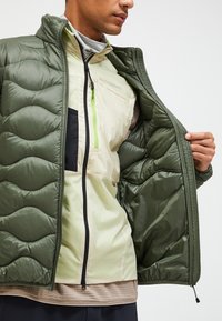 Chaqueta acolchada de color verde oliva con textura suave, llevada sobre una capa interior de color amarillo claro con acentos negros y un cierre frontal con cremallera.