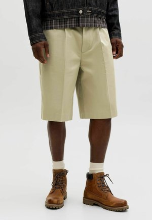 Personne portant un short beige jusqu'aux genoux, des chaussettes blanches, des bottes marron à lacets et une veste sombre sur un fond clair uni.