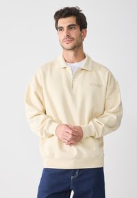 Sudadera de color crema con cremallera de un cuarto, puños acanalados y un ajuste relajado. Presenta el texto "KYOTO, JAPÓN" bordado en el pecho.