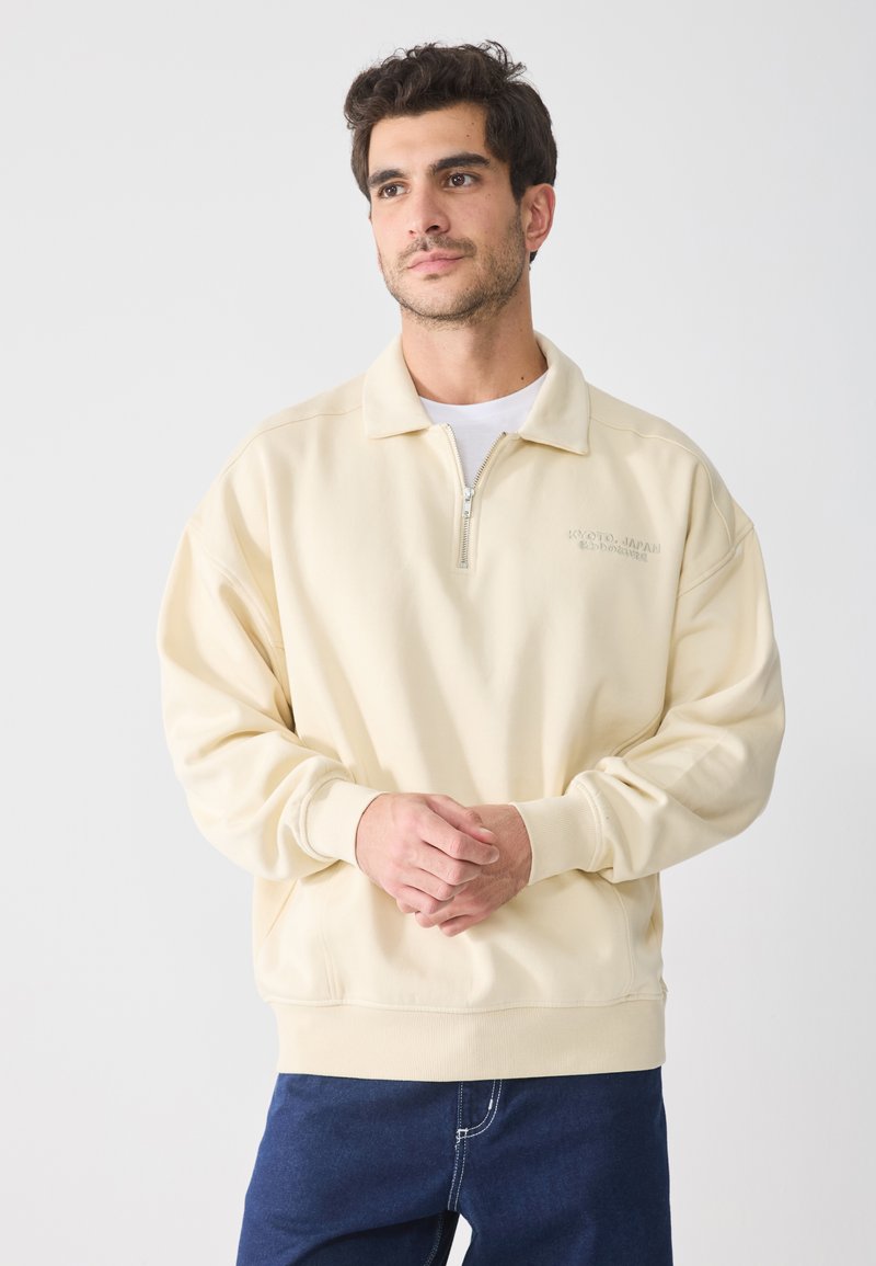 Sudadera de color crema con cremallera de un cuarto, puños acanalados y un ajuste relajado. Presenta el texto "KYOTO, JAPÓN" bordado en el pecho.