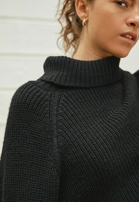 Woman wearing black knitted turtleneck sweater and gold hoop earrings with curly hair pulled back against a light background.