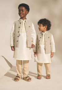 Deux garçons portent des tenues traditionnelles : des kurtas crème avec des gilets beige, des détails brodés et des pantalons clairs. Ils portent des sandales beige.