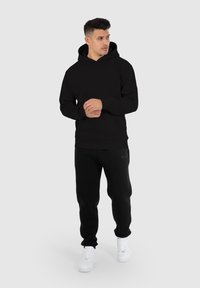 Sudadera con capucha negra hecha de tela suave, con un bolsillo en la parte frontal y puños de canalé. Combinada con pantalones de chándal negros y zapatillas blancas.