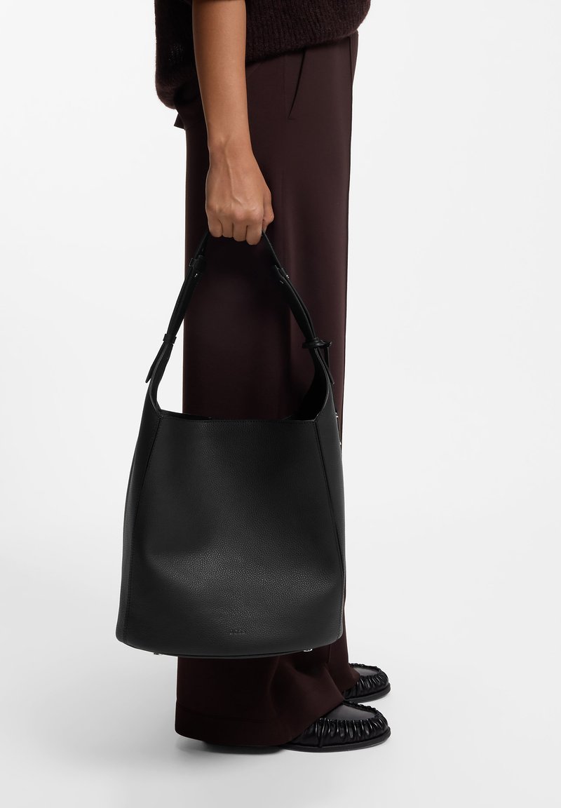 Personne tenant un grand sac à main en cuir noir par sa poignée à côté d'un pantalon large marron foncé et de chaussures en cuir noires froncées.