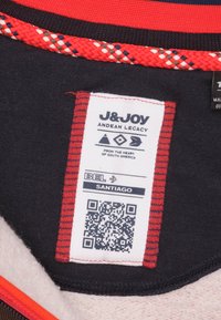 Zwarte stof met rode gestreepte accenten en een geweven label met een QR-code. Het label bevat de merknaam "J&JOY" en informatie over het land van oorsprong.