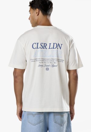 Homme portant une chemise blanche à manches courtes avec texte bleu "CLSR LDN ATELIER" et une petite impression supplémentaire dans le dos, associée à un jean bleu clair.