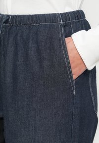 Tamsiai mėlynos spalvos džinso kelnės su elastine liemens dalimi, šoninėmis kišenėmis ir baltais siūlų detalizacijomis. Artimas audinio tekstūros vaizdas.