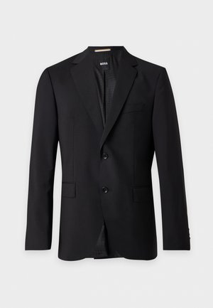 Un homme se tient devant un fond gris clair, vêtu d’un blazer noir, d’une chemise blanche, d’un pantalon taupe et de mocassins noirs.