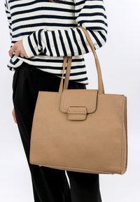 Sac à main en cuir texturé beige avec une fermeture à rabat, deux poignées et un logo discret. Une femme portant une chemise rayée tient le sac.