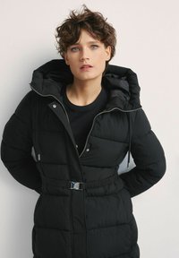 Czarna kurtka puchowa z kapturem, zapinana na zamek z przodu i z wąskim krojem w talii. Posiada pikowane sekcje oraz srebrne akcenty.