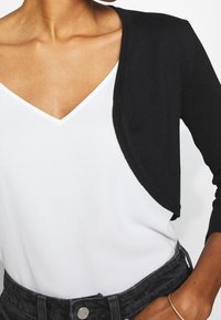 Musta lyhyt neuletakki valkoisen v-muotoisen yläosan päällä. Neuletakissa on pehmeä pinta ja avoin etuosa, pitkät hihat ja hillitty koristelu.