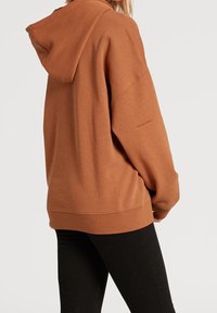 Sweat à capuche brun avec cordon de serrage, coupe oversize, ourlet et poignets côtelés, avec un logo sur la manche. Leggings noirs visibles.