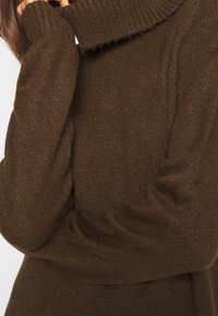 Maglione a collo alto in maglia marrone con maniche lunghe, vestibilità leggermente oversize, indossato da una persona con una mano parzialmente visibile vicino al viso.