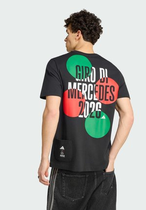 Man in zwart t-shirt met grote rode en groene cirkels en de tekst "Giro di Mercedes 2026" op de rug, draagt donkere jeans met gestreepte zijkanten.