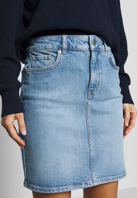 Helles blaues Denim-Rock mit taillierter Passform, der Fronttaschen, Gürtelschlaufen und sichtbare Nähte entlang des Saums und der Nähte aufweist.