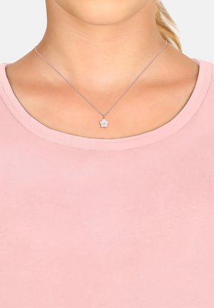 Sølv halskæde med en lille blomstervedhæng, der har en central ædelsten, båret over en lys pink tætsiddende top med en rund halsudskæring.