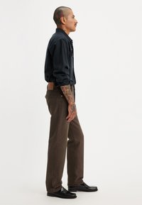 Camisa abotonada azul oscuro, pantalones marrones y zapatos negros brillantes. Tatuajes notables en los brazos, textura de tela rugosa y corte clásico.
