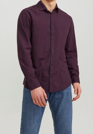 Mand iført en langærmet, mørkerød og marineblå ternet skjorte med blå jeans, står mod en ensfarvet lys baggrund.