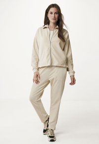 Beige zip-up jacka med krage och matchande tapered byxor. Båda har en texturerad yta. Bärs med svarta och beige sneakers.