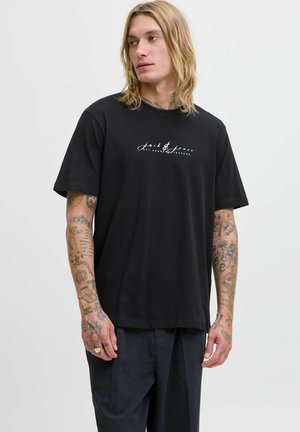 Bărbat cu păr blond până la umeri și tatuaje pe brațe, îmbrăcat cu un tricou negru cu text alb și pantaloni închiși la culoare, stând în fața unui fundal simplu.