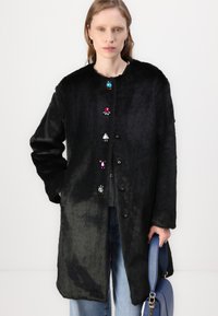 Črn plašč iz umetnega krzna s krožnim ovratnikom, okrašen s pisano podobnimi dragulji gumbi navzdol po sprednji strani. Tkanina ima mehko teksturo.