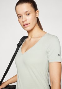 Svijetlozeleni T-shirt s V-izrezom izrađen od mekane tkanine, kratkih rukava, s malim crnim logotipom na lijevom rukavu. Nosim torbu s crnim remenom.