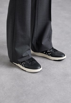 Baskets en cuir noir avec semelle en gomme blanche, présentant des lacets et des accents géométriques. Associées à un pantalon large gris.