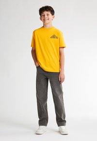 Camiseta de algod�ón amarilla con un gráfico negro impreso, combinada con pantalones de pana gris y zapatillas blancas. El modelo está de pie con una mano en el bolsillo.
