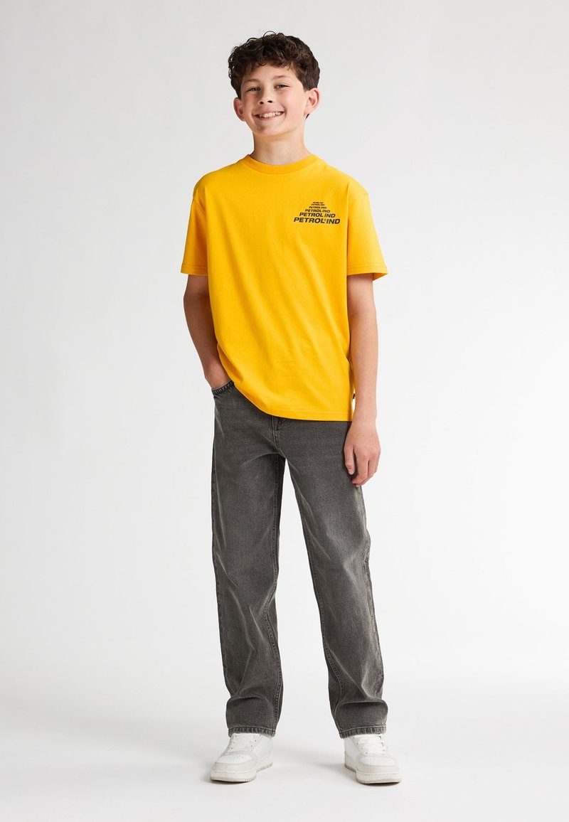 Camiseta de algodón amarilla con un gráfico negro impreso, combinada con pantalones de pana gris y zapatillas blancas. El modelo está de pie con una mano en el bolsillo.