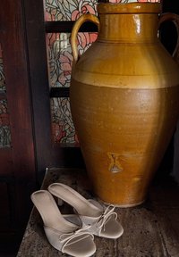 Beiga öppna-toe klackar med en texturerad överdel och en rosettdetalj, placerade bredvid en stor brun keramisk vas med två handtag.