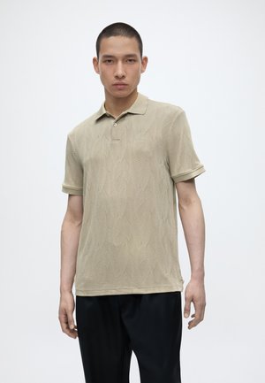 Jeune homme aux cheveux courts porte un polo beige texturé et un pantalon noir, debout devant un fond clair uni.