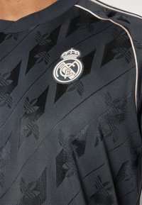 Jersey atlético negro con patrones de diamante texturizados y un logo blanco del Real Madrid en el pecho, con una franja clara en el hombro.