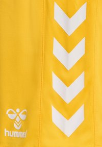 Gul idrottskläder med vita chevronmönster och "hummel"-logotypen längst ner. Tillverkad av ett strukturerat och hållbart tyg.