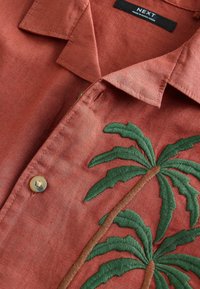 Chemise en lin de couleur rouille avec un motif de palmier brodé en vert et marron. Comprend un détail de bouton près du col.