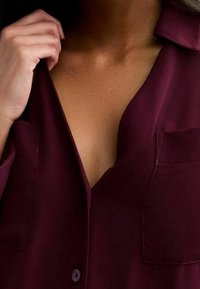 Burgundy button-up shirt with a collar, two front pockets, and a V-neckline. Smooth texture with a slightly loose fit.