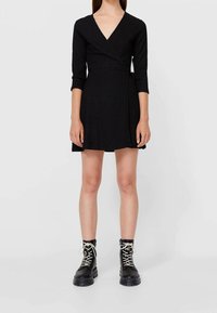 Robe portefeuille noire à décolleté en V profond, manches longues et tissu texturé, associée à des bottes de combat noires à lacets avec une semelle épaisse.