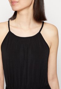 Femme portant une robe noire sans manches avec des fines bretelles, montrant son cou, ses épaules et une partie de son visage tourné légèrement à gauche.