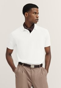 Weißes Polohemd mit kurzen Ärmeln, strukturiertem Muster und V-Ausschnitt, kombiniert mit braunen Hosen und einem passenden braunen Schal.