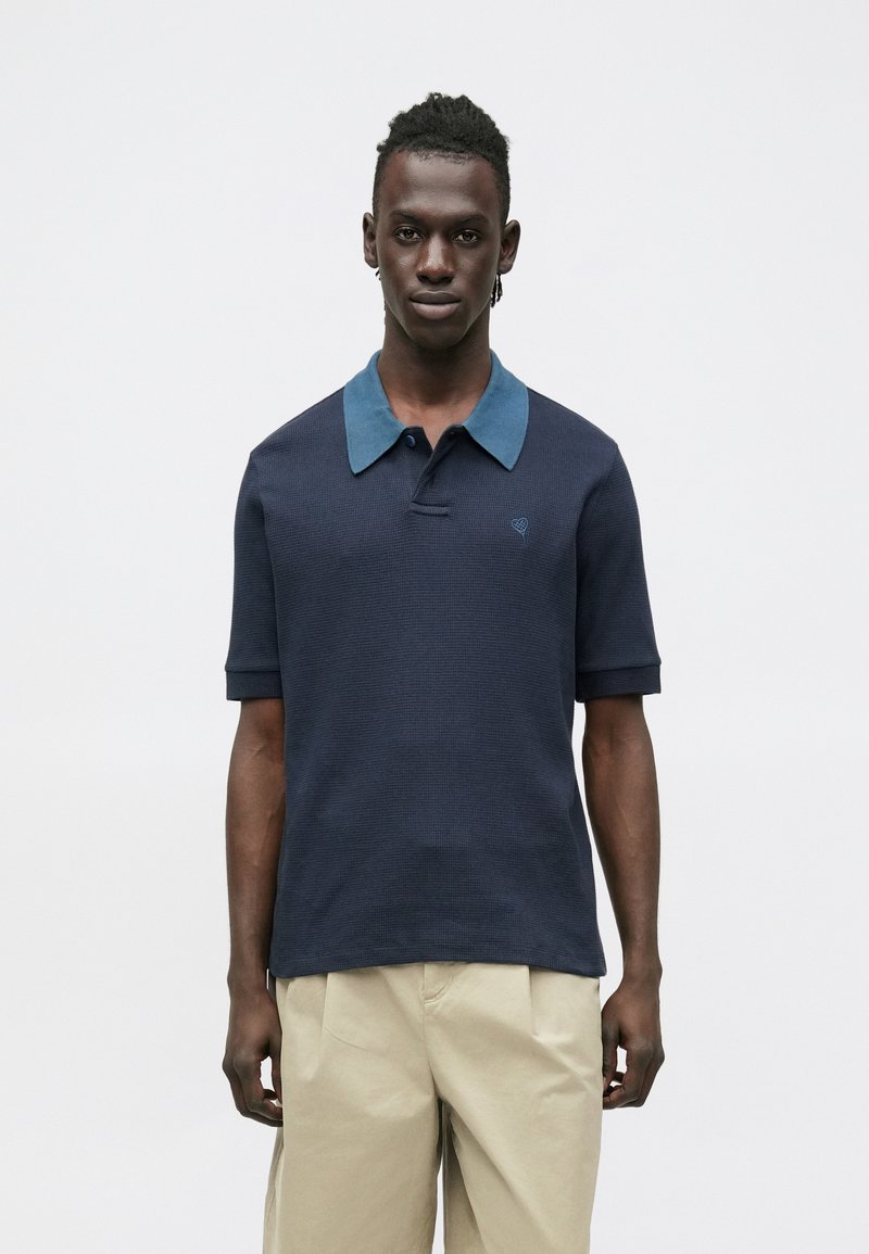 Tmavomodré polo tričko z textúrovaného materiálu s kontrastným svetlomodrým golierom a malým vyšívaným logom na hrudi.