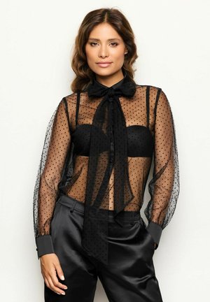 Black sheer blouse with polka dots, a bow tie at the collar, and long sleeves. Worn over a black strapless top and paired with satin trousers.