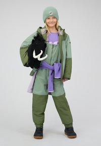 Grüne Winterjacke über einem lila Outfit, hält ein schwarzes Mammut-Plüschtier. Enthält Kunstlederstiefel und eine hellgrüne Mütze.